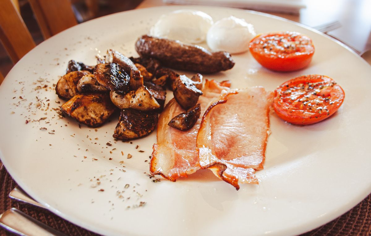 A cooked breakfast plate with sausage, back bacon, sautéed mushrooms, poached eggs and grilled tomatoes