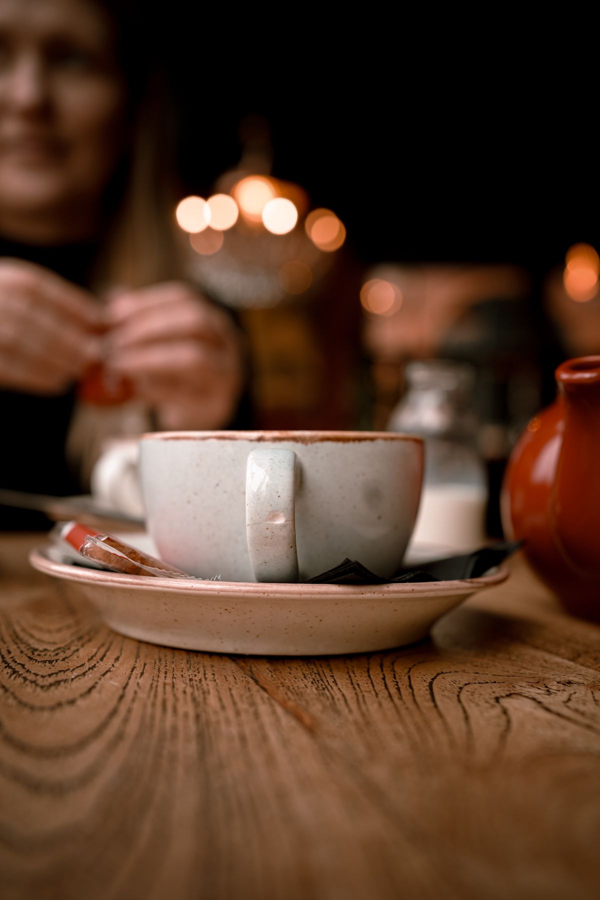 A speckled ceramic mug and saucer with a small jug of milk on a warm wooden café table